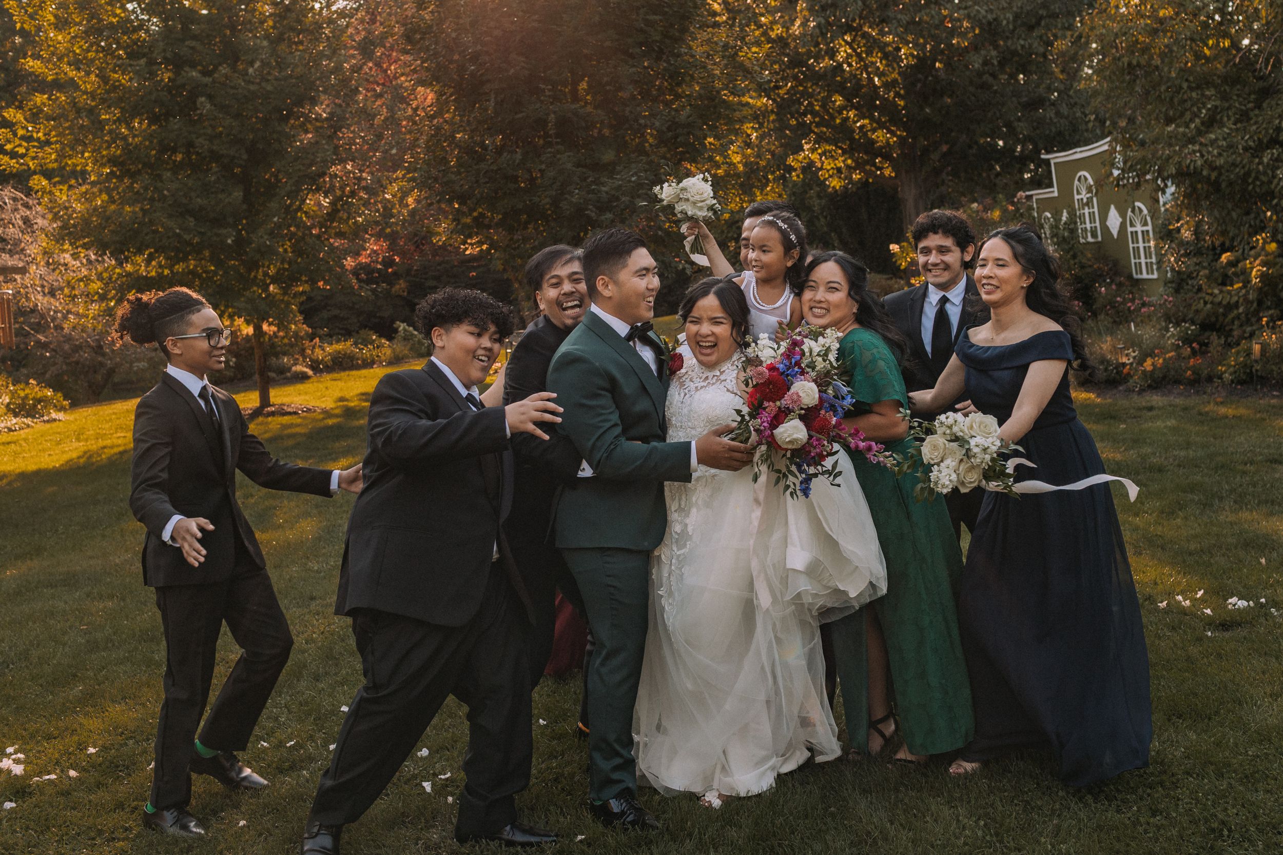 Wedding party members in dark suits and green dresses laugh together in a fall garden setting with golden foliage.