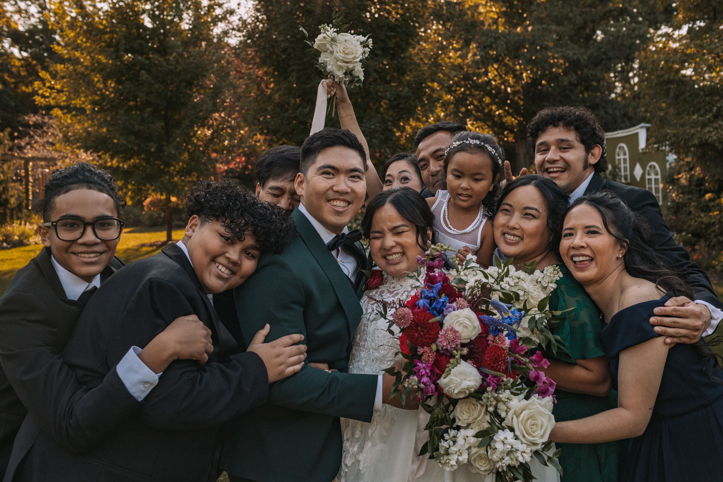 Wedding party celebrates outdoors during golden hour in autumn with colorful fall foliage in background.
