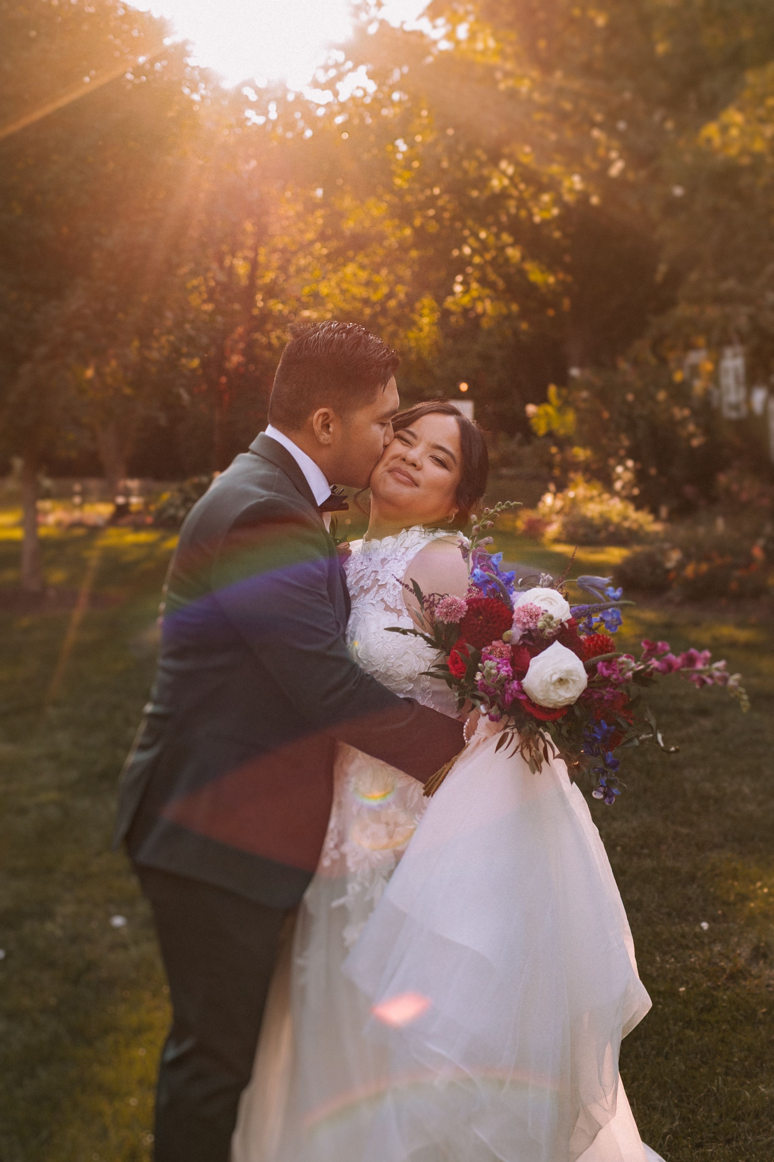 A romantic wedding couple embraces in a garden at golden hour with sunlight streaming through autumn trees.