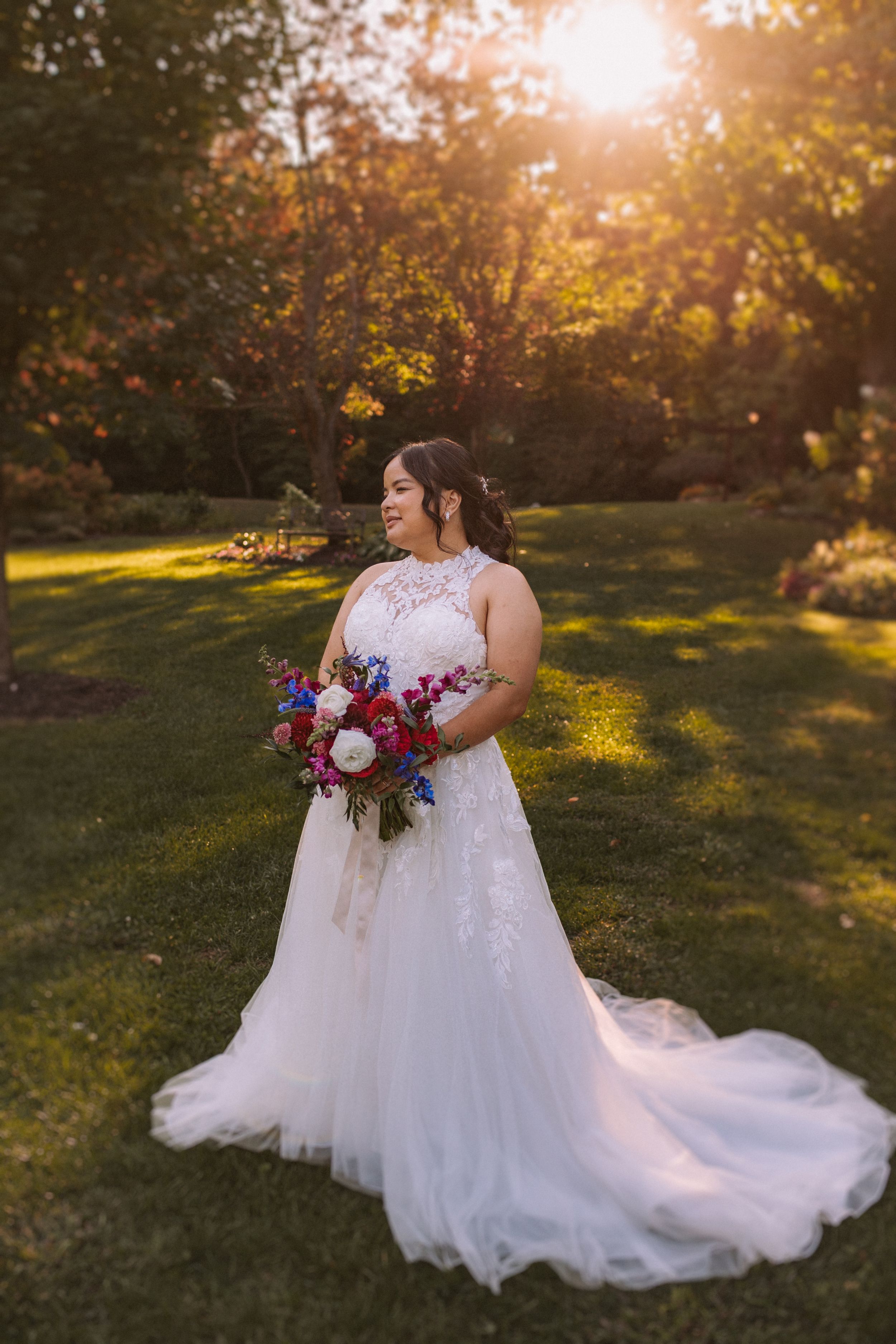 A bride in a flowing white gown poses in a garden at sunset with a burgundy and pink bouquet.