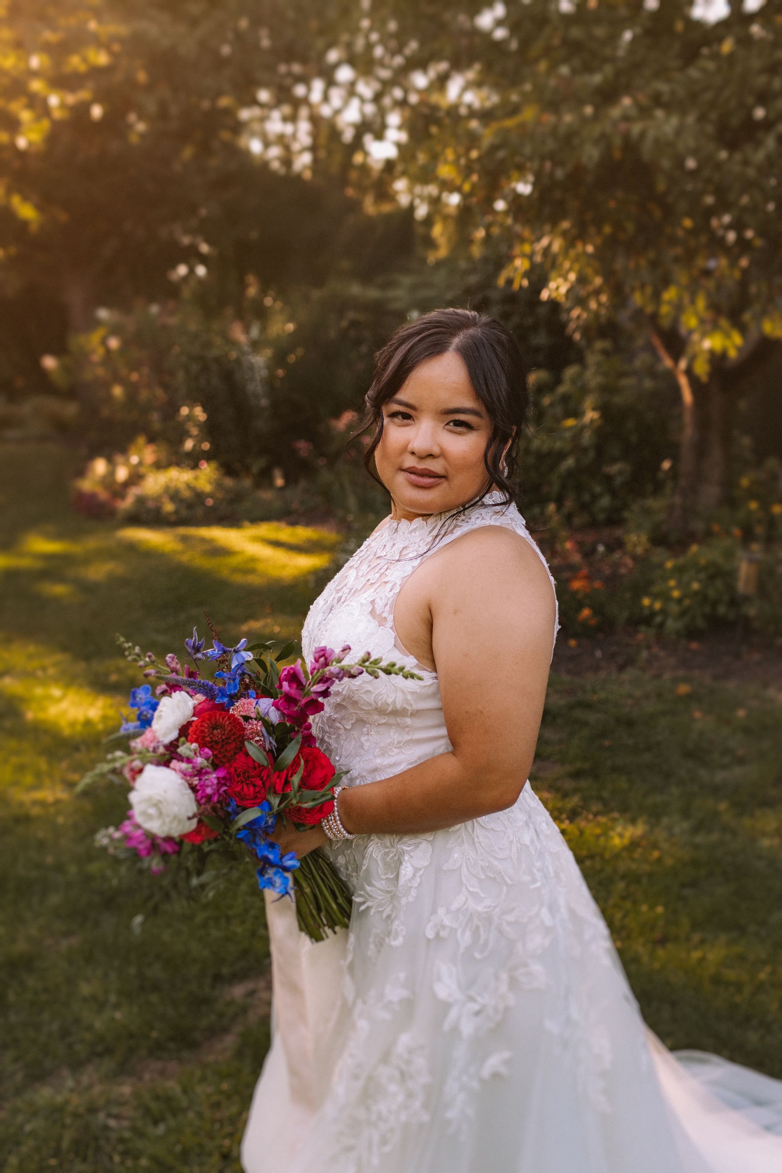 A beautiful wedding bouquet with vibrant red and blue flowers held against a white lace dress in golden sunlight.