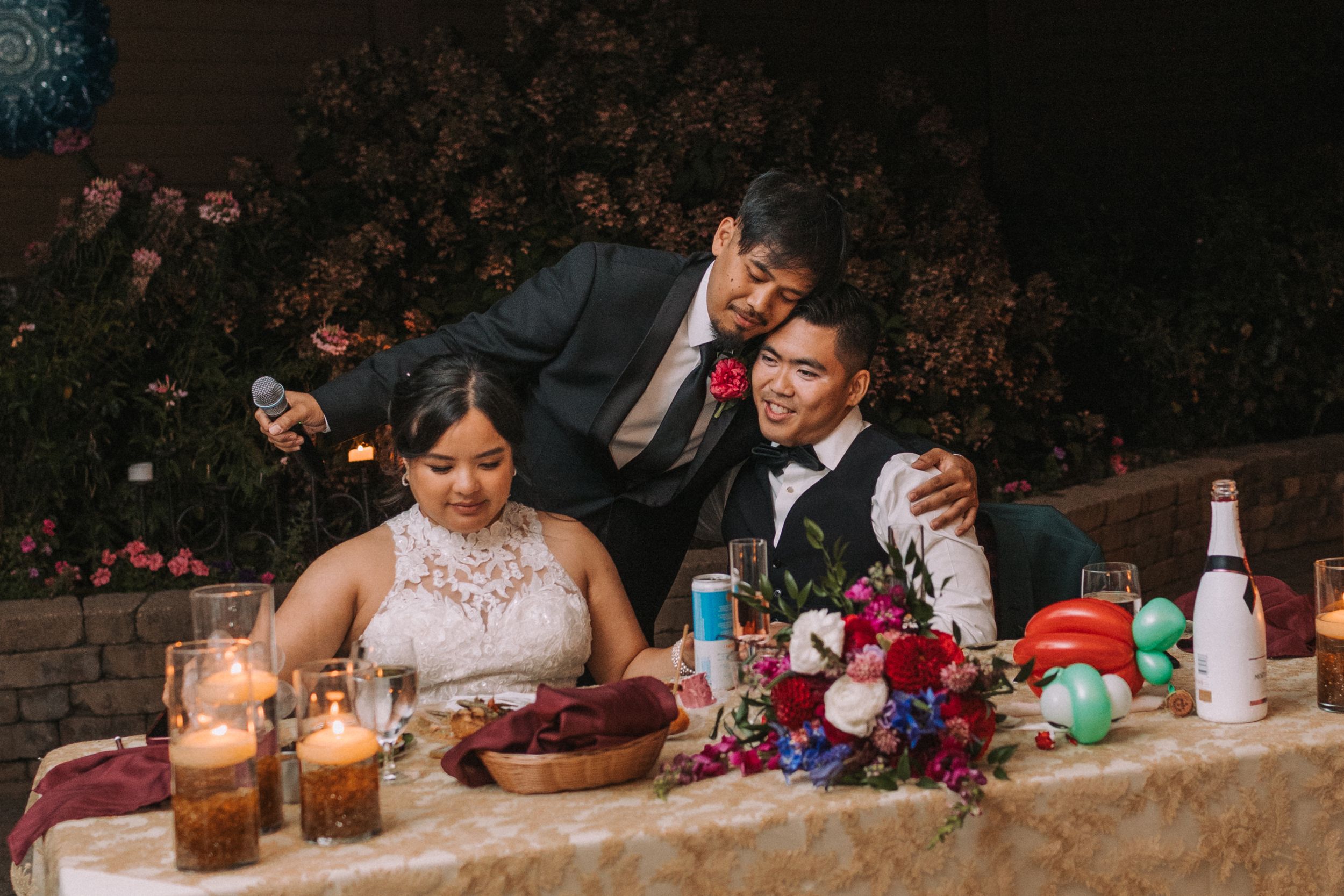 Wedding reception guests sit at a long table during a dimly lit evening celebration with drinks and floral decor.