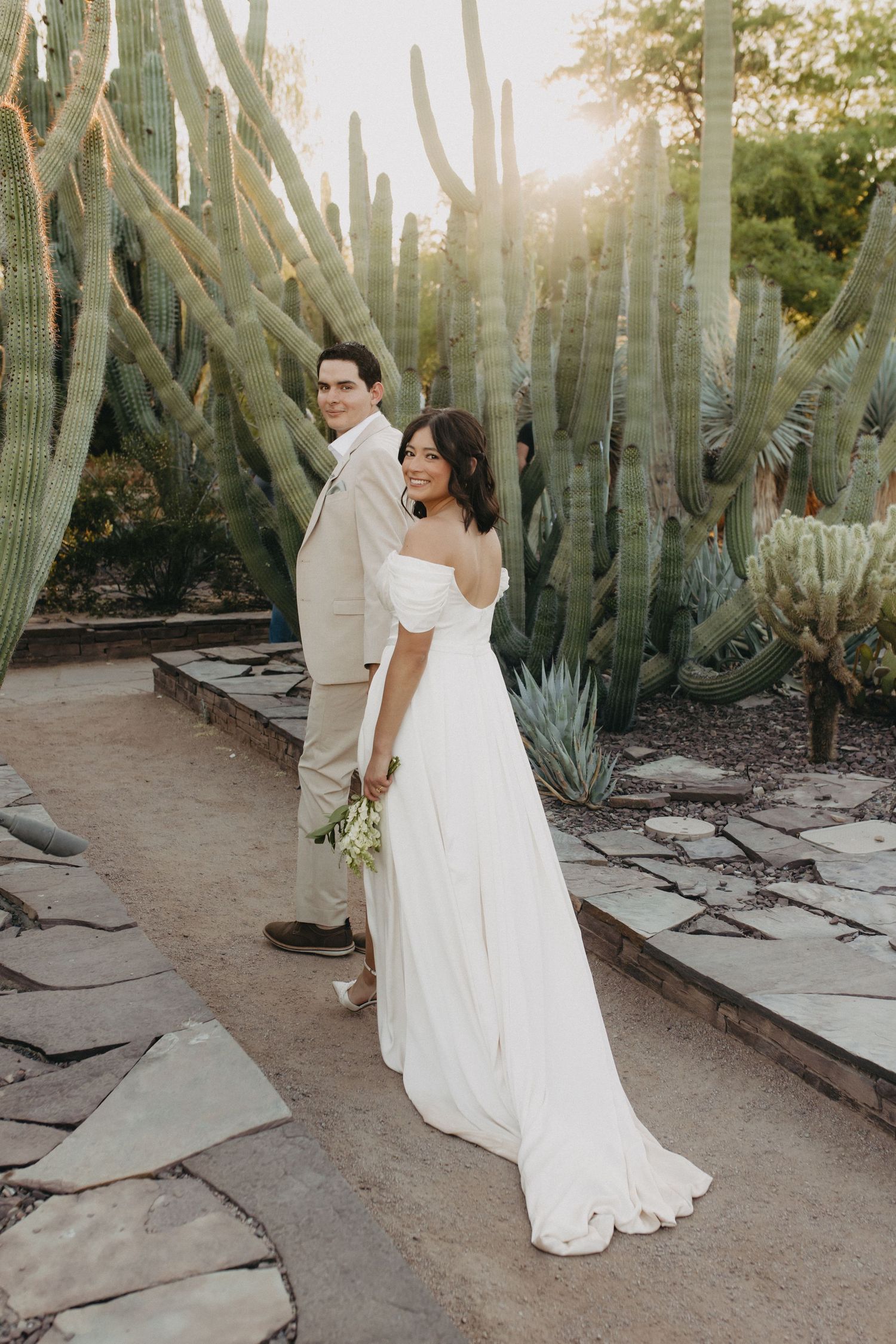 A couple in wedding attire embraces while standing among tall desert cactus at sunset.