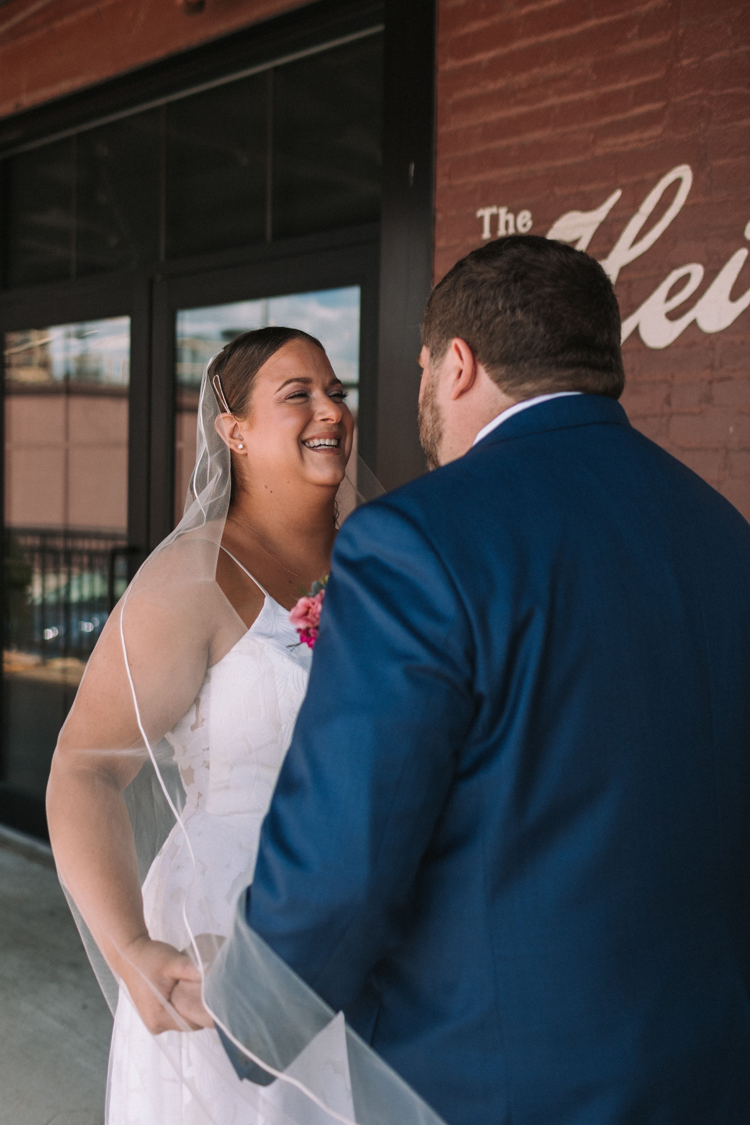 A bride in white dress shares a joyful moment with groom in navy suit during their first look outside a brick building.