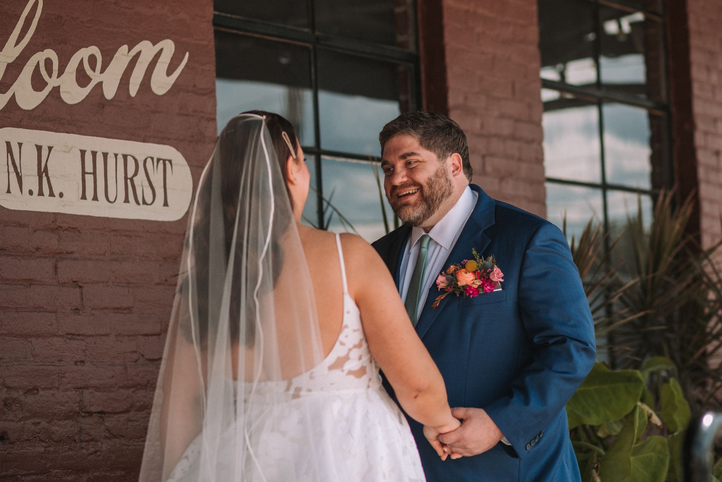 Elegant couple in wedding attire standing against a rustic brick wall with vintage signage.
