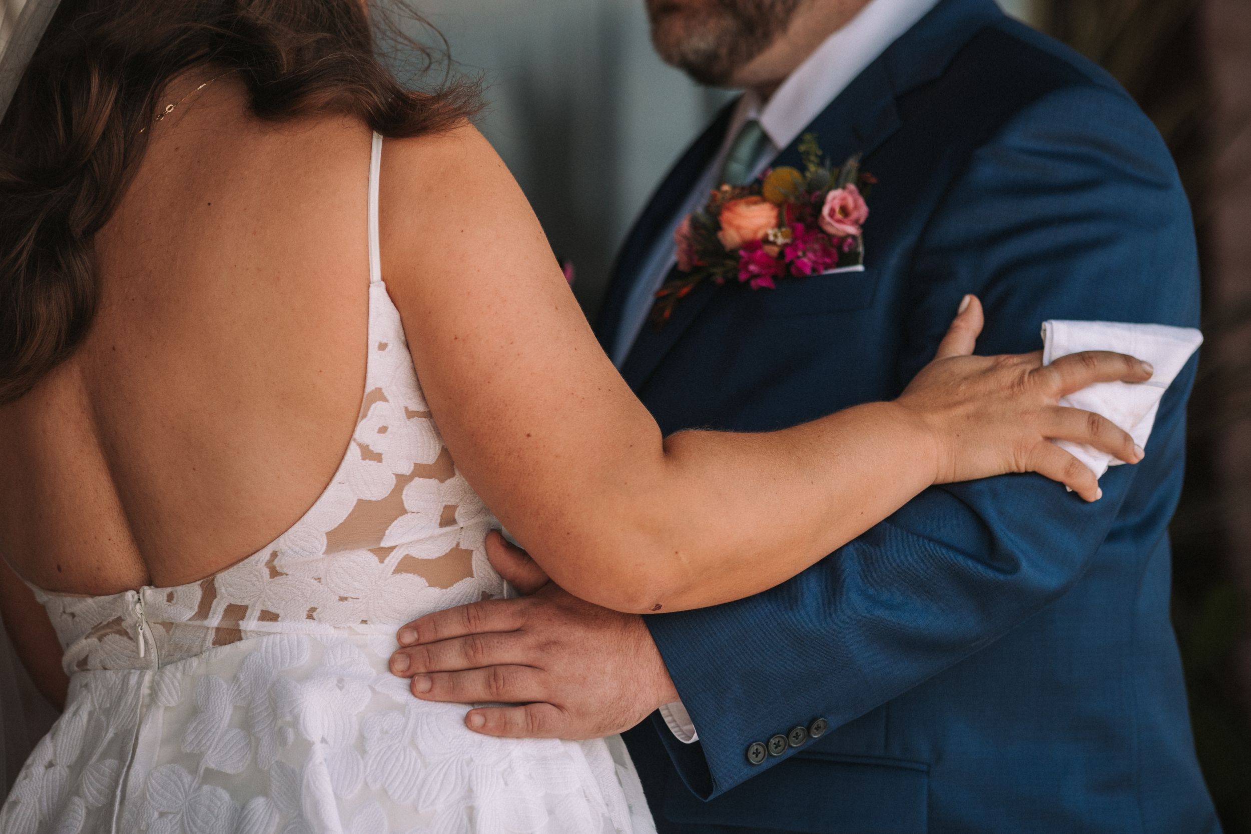 Close-up details of a couple embracing in wedding attire, showing the delicate lace of the bridal gown and navy suit.