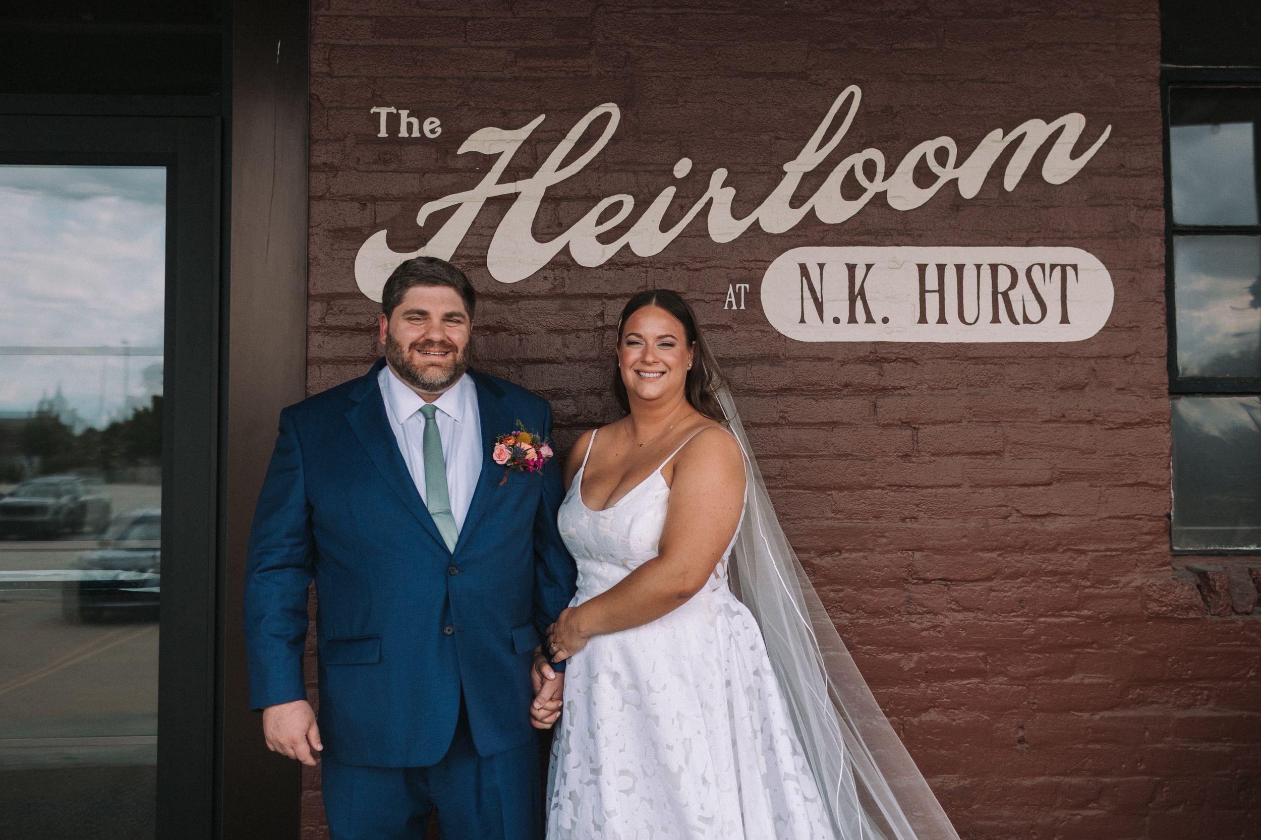 Wedding couple posing in front of a red brick wall with 'Heirloom' and 'N.K. Hurst' signage.