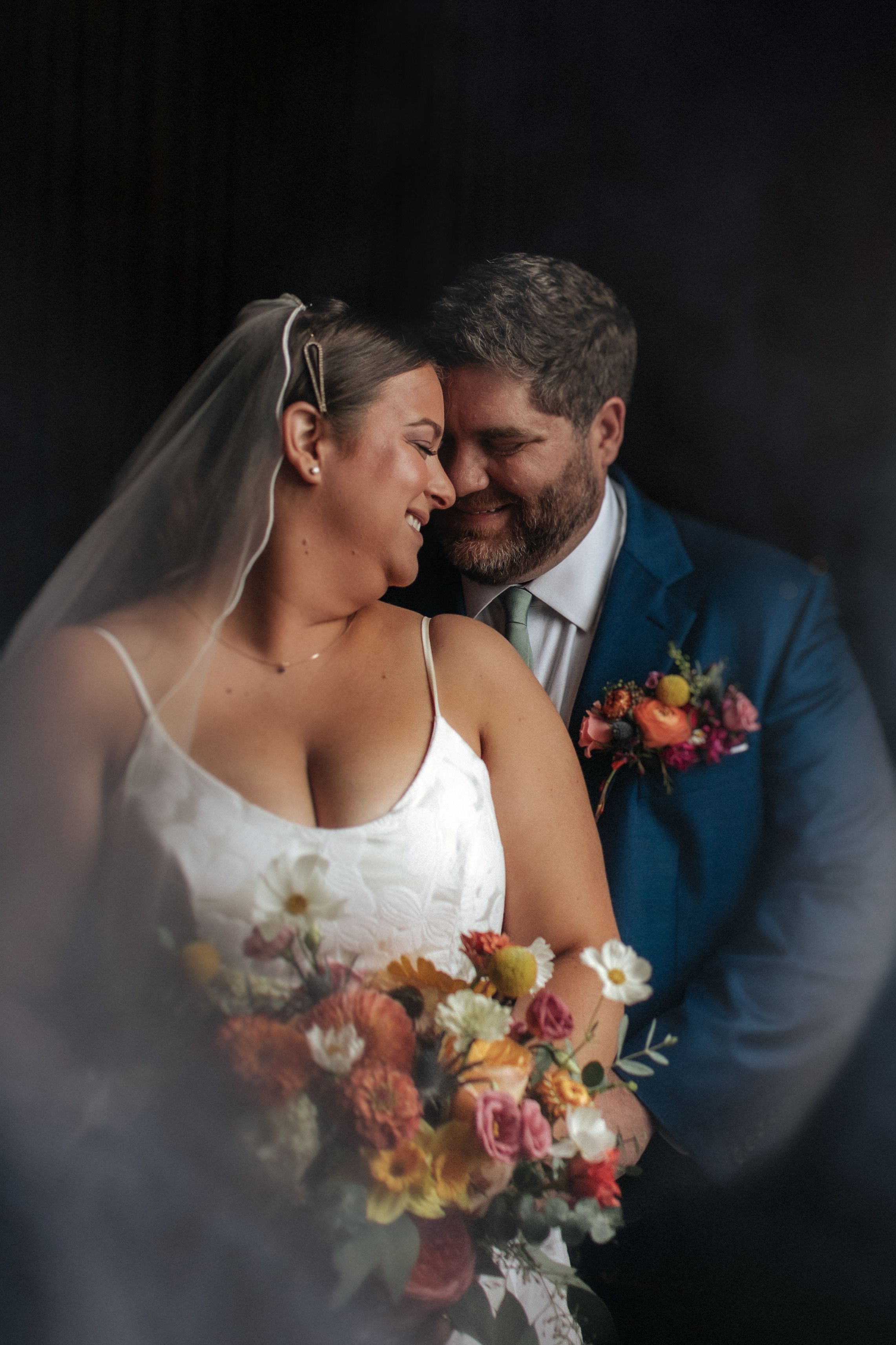 Intimate wedding portrait series showing couple embracing with colorful floral bouquet against dark background.