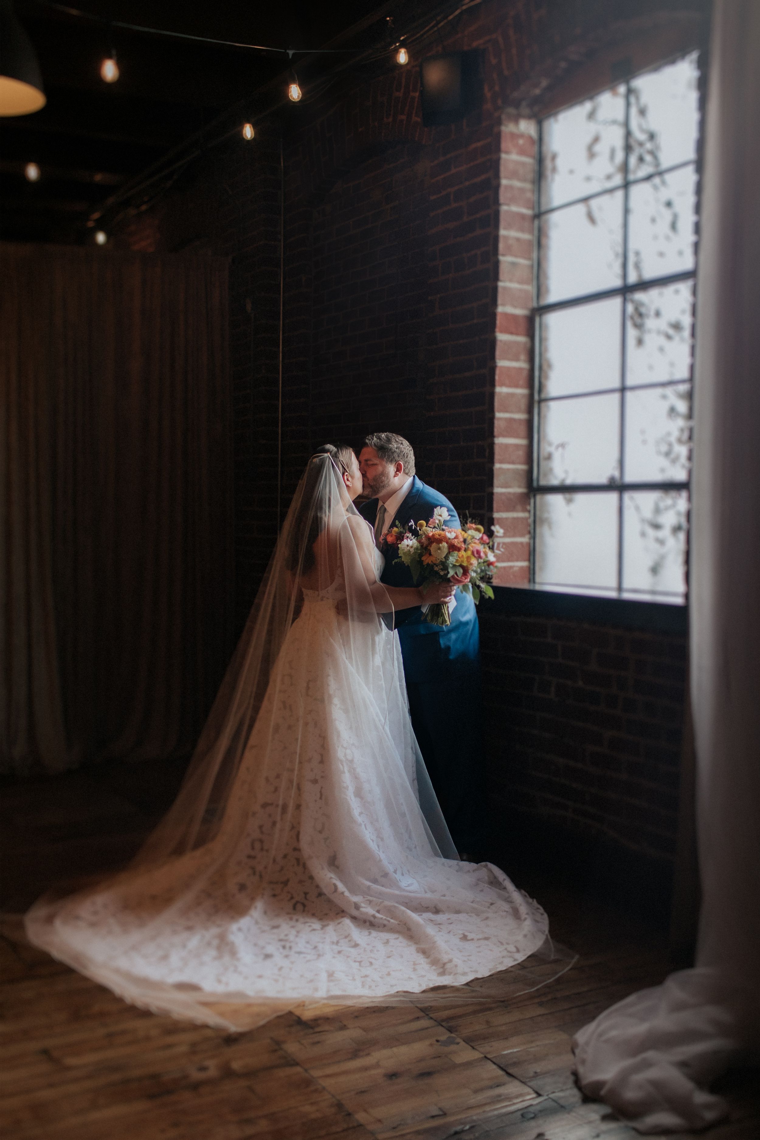 A bride in a flowing white wedding gown with a long cathedral veil sits on a rustic wooden floor near large windows.