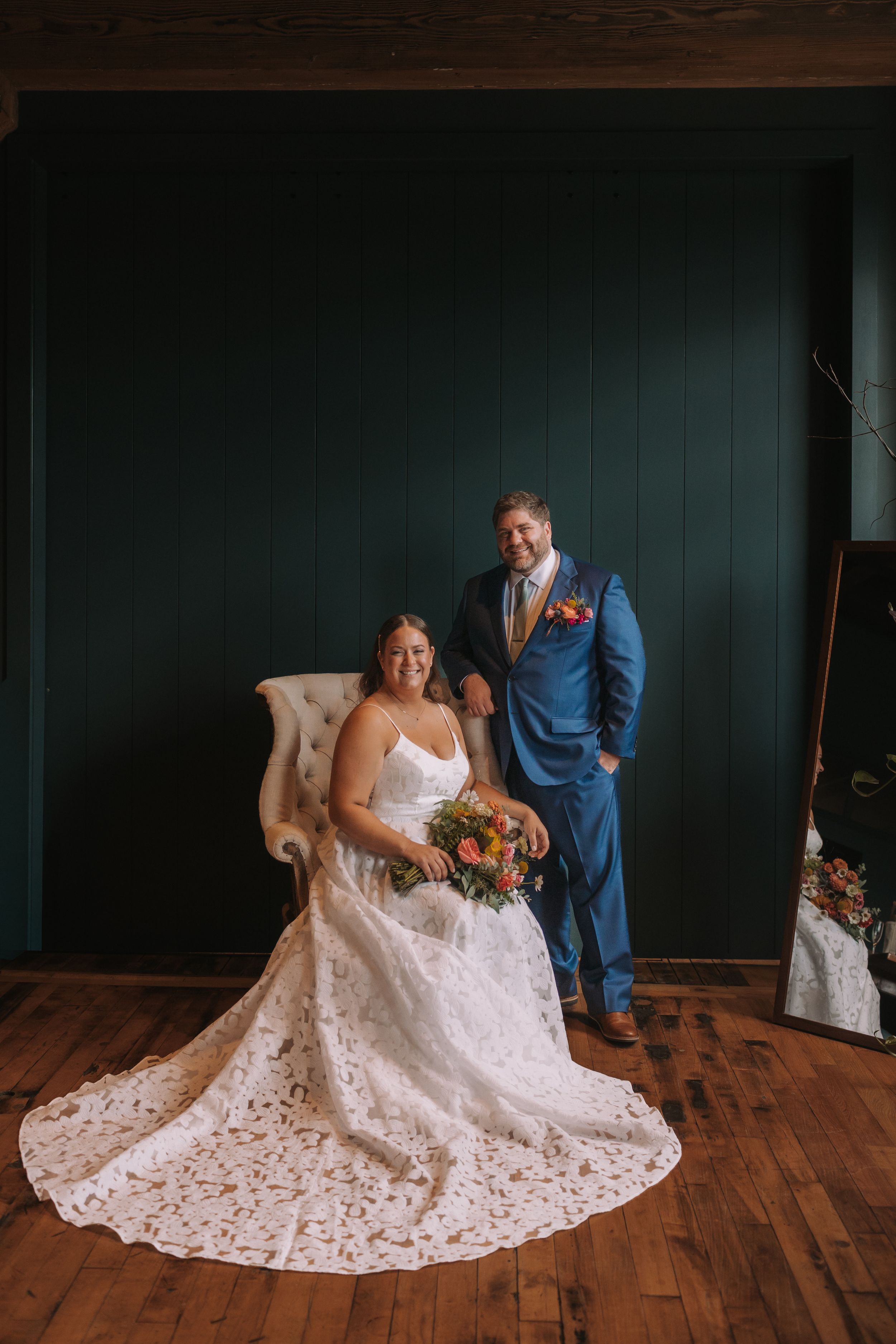 A romantic wedding scene shows a couple in formal attire posing together on wooden floors against a dark teal wall.
