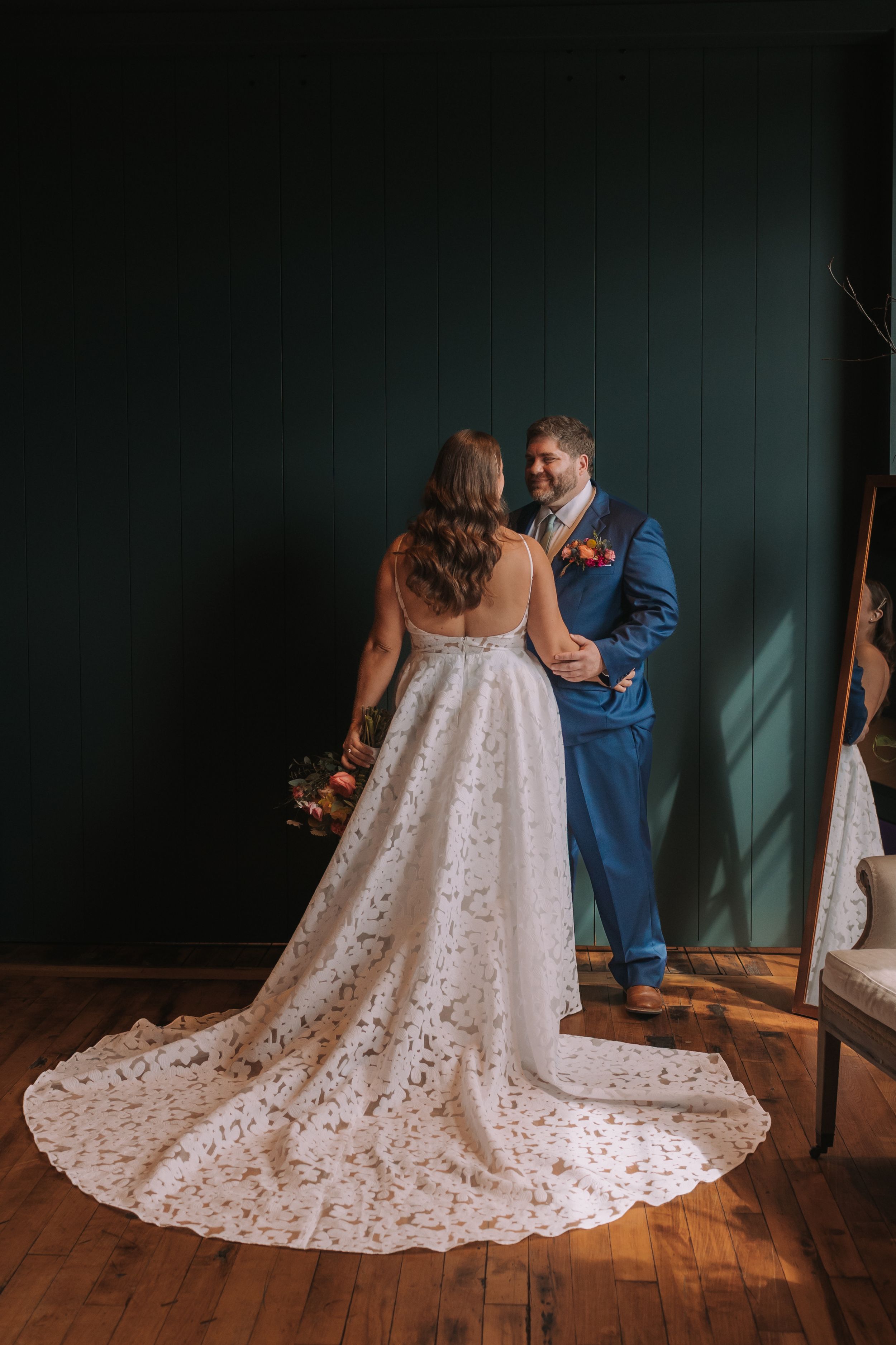 A wedding dress with intricate lace details and train displayed in multiple angles against a dark green wall and wooden floor.