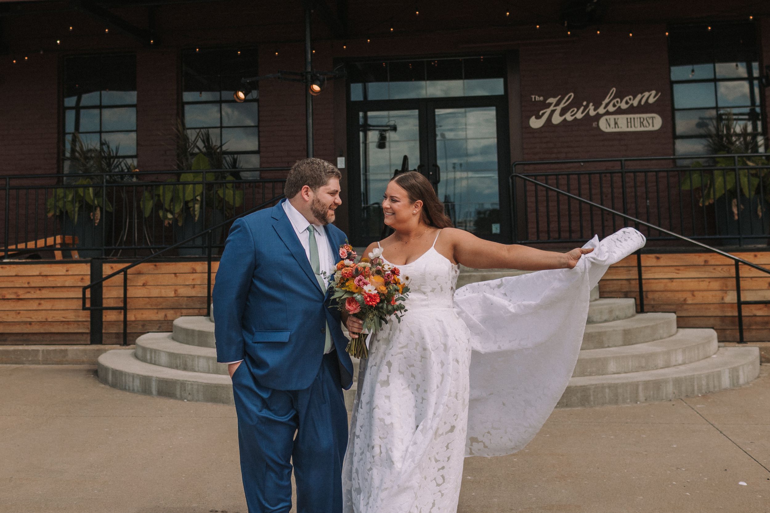 A joyful couple shares a romantic moment on stone steps outside a brick building with their wedding attire and bouquet.