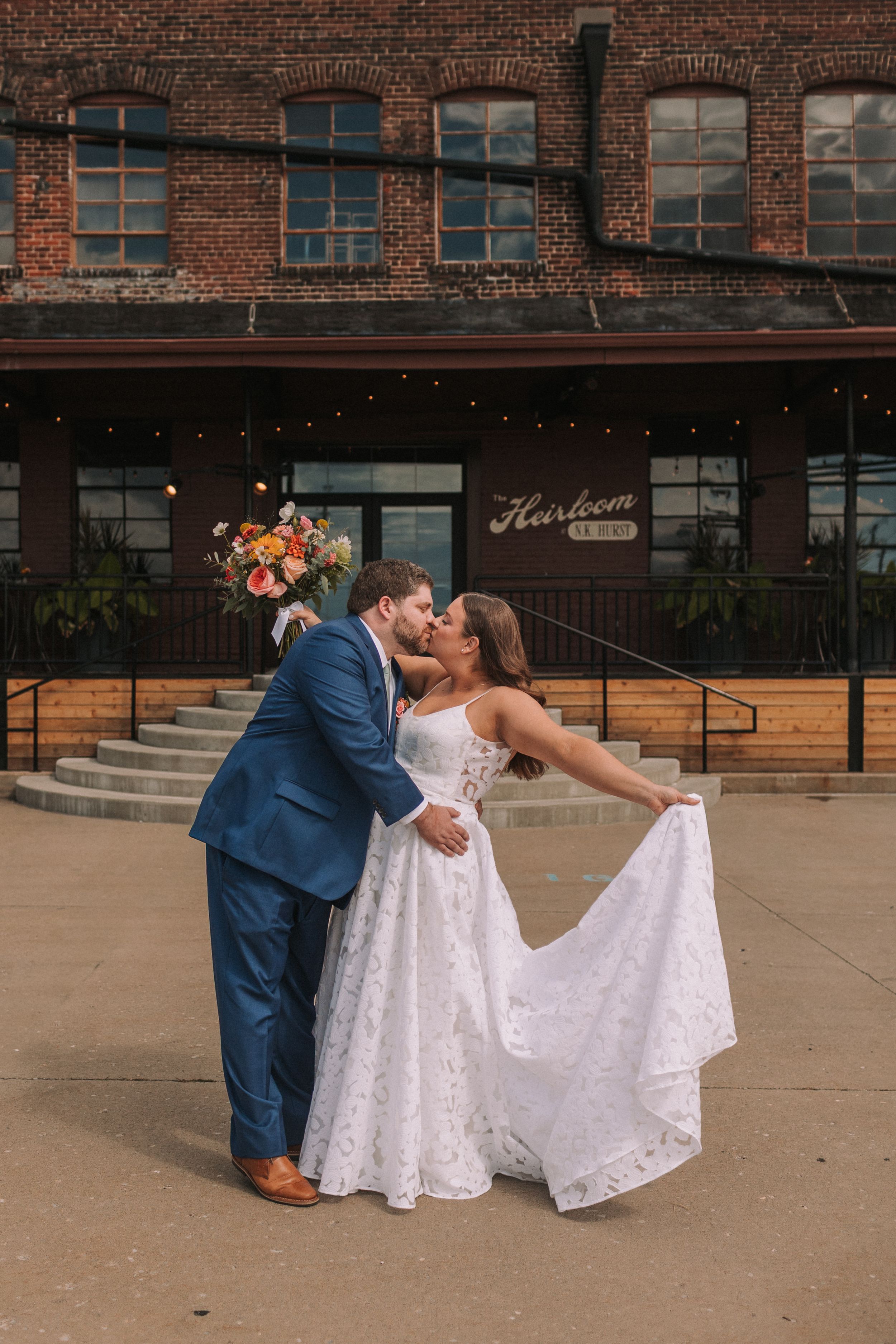 A romantic couple in formal attire dips and poses together in front of a rustic brick building with string lights.