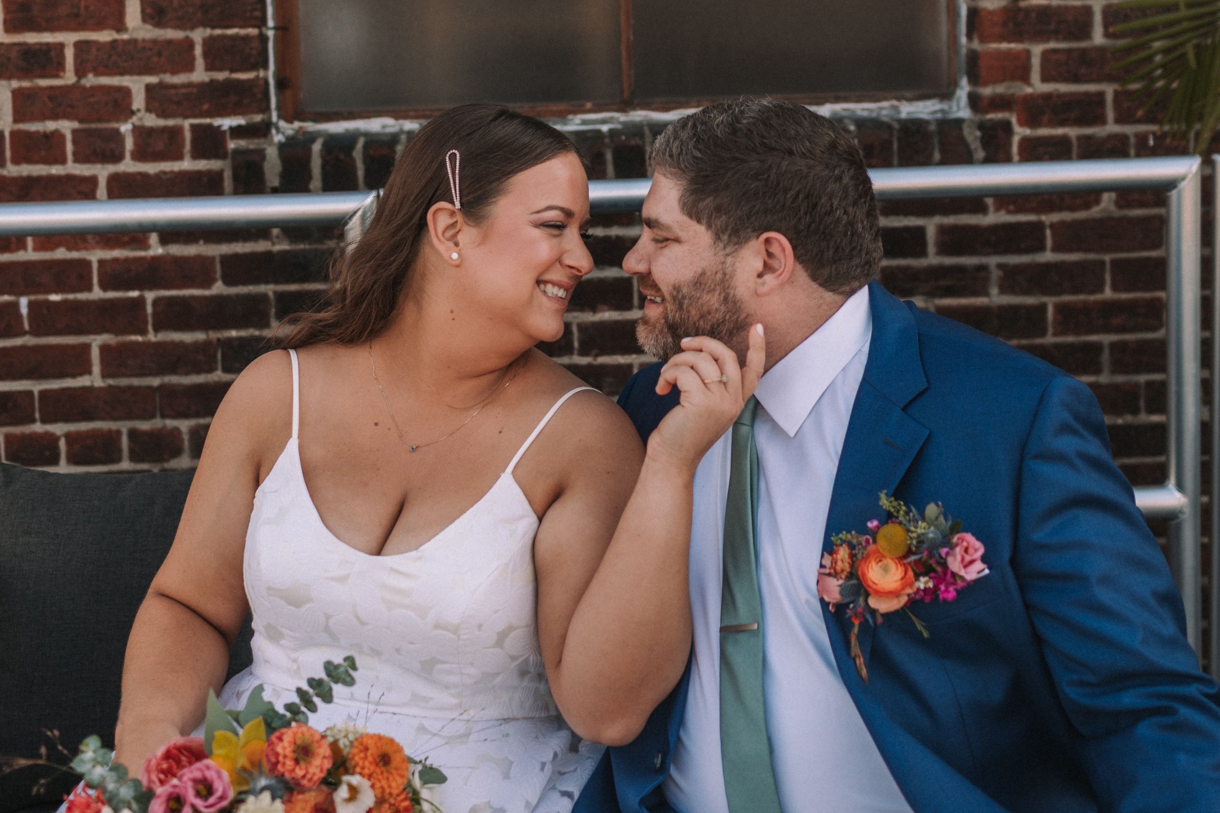 A happy couple shares an intimate laugh while holding a colorful floral bouquet against a brick wall backdrop.
