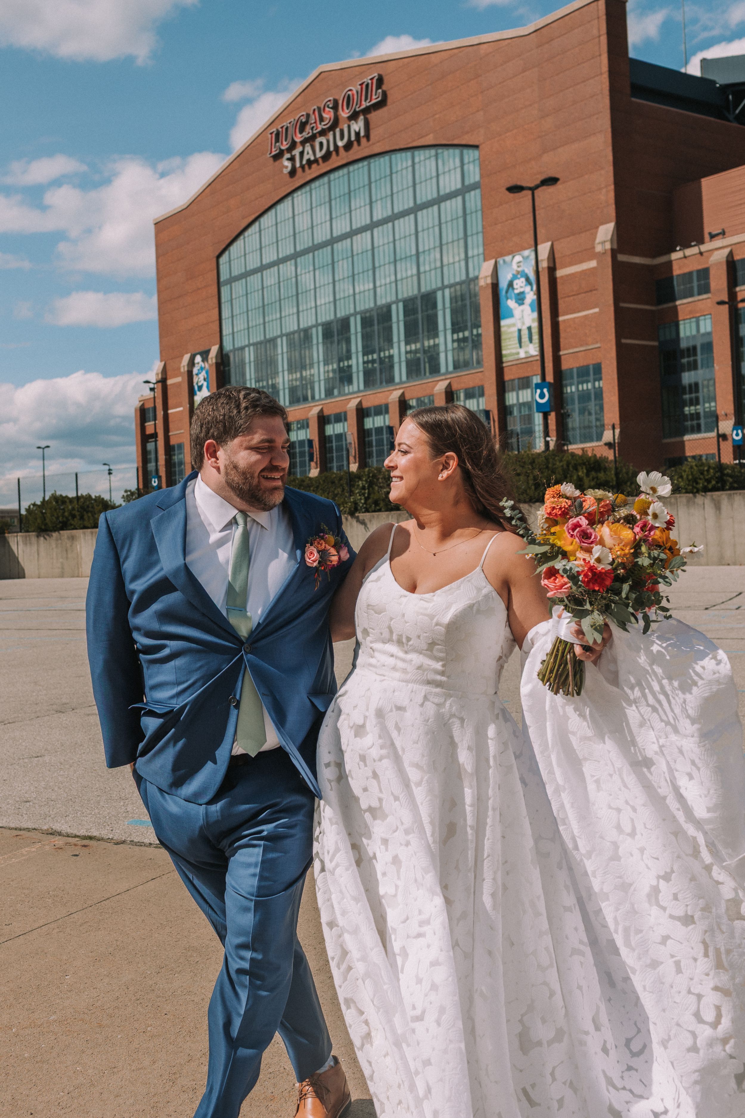 A couple in wedding attire share a joyful moment outside a modern brick and glass building on a sunny day.