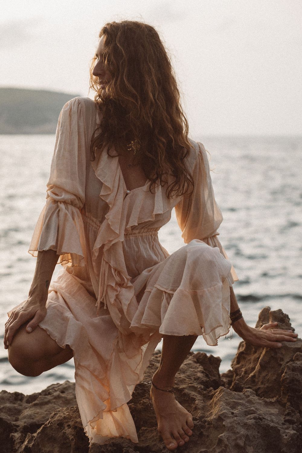Woman in flowing white dress sits on rocky coastal shore at sunset with ocean waves in background.