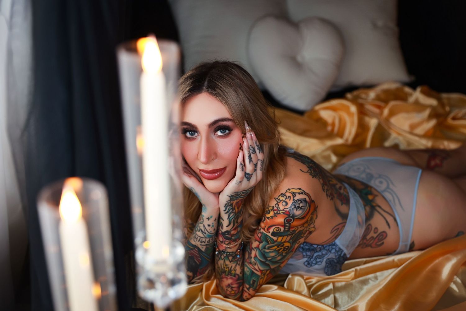 Candlelit boudoir photo featuring tattoos and golden satin sheets in moody lighting.