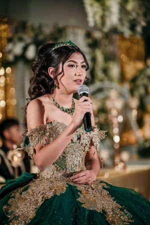 A person in an emerald green and gold quinceañera gown speaks into a microphone during a celebration.
