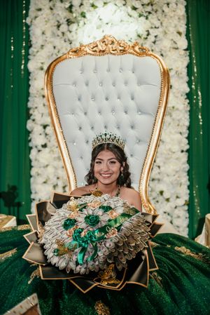 A celebrant sits on an ornate white and gold throne chair holding a luxurious green and gold bouquet.