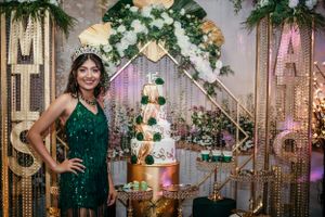 Person in emerald sequin dress poses beside elegant tiered white cake and gold decor.
