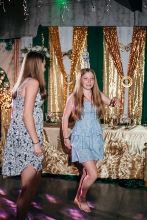 Two young people in casual dresses dance at a party with gold and green decorations.