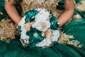 A bouquet featuring white and teal roses with gold accents rests on emerald fabric.