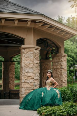An elegant green ball gown photographed under a stone gazebo in a lush garden setting.