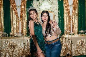 Two people pose together in front of a sparkly gold and green backdrop at an event.