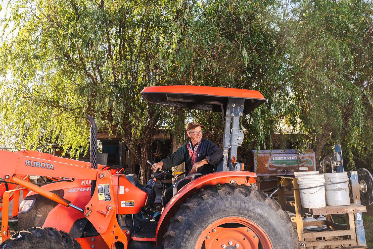 Orange Kubota tractor parked near trees and equipment in an outdoor work area.