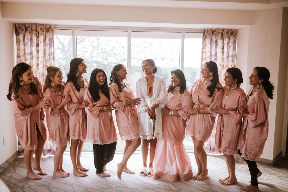 A group in matching blush pink satin robes gather by a window for a pre-wedding celebration moment.