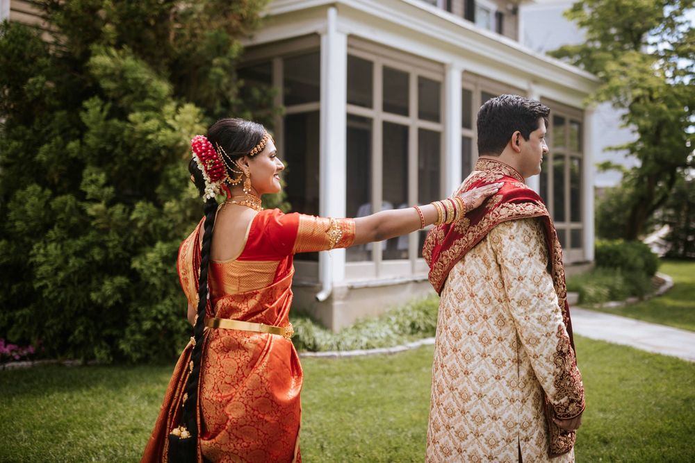 Two people in traditional Indian wedding attire share a joyful moment on a green lawn outside a white building.