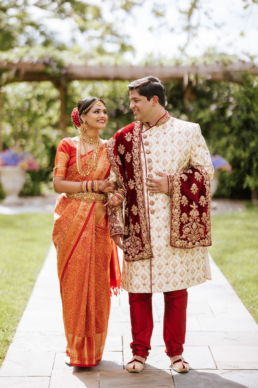 Traditional Indian wedding couple in ornate red and cream outfits pose together on garden pathway.