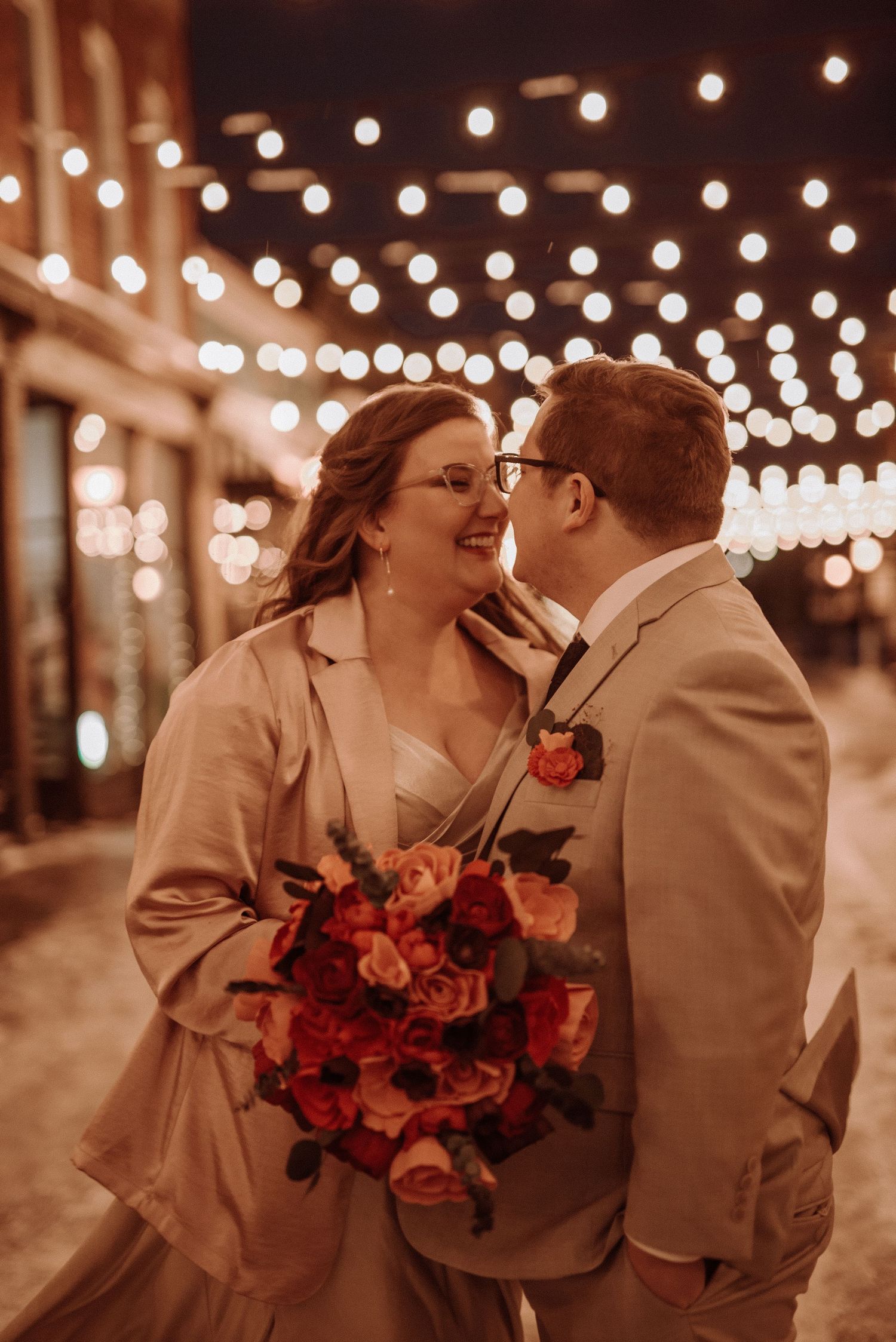 A romantic wedding couple shares an intimate moment under glowing string lights while holding a vibrant red bouquet.