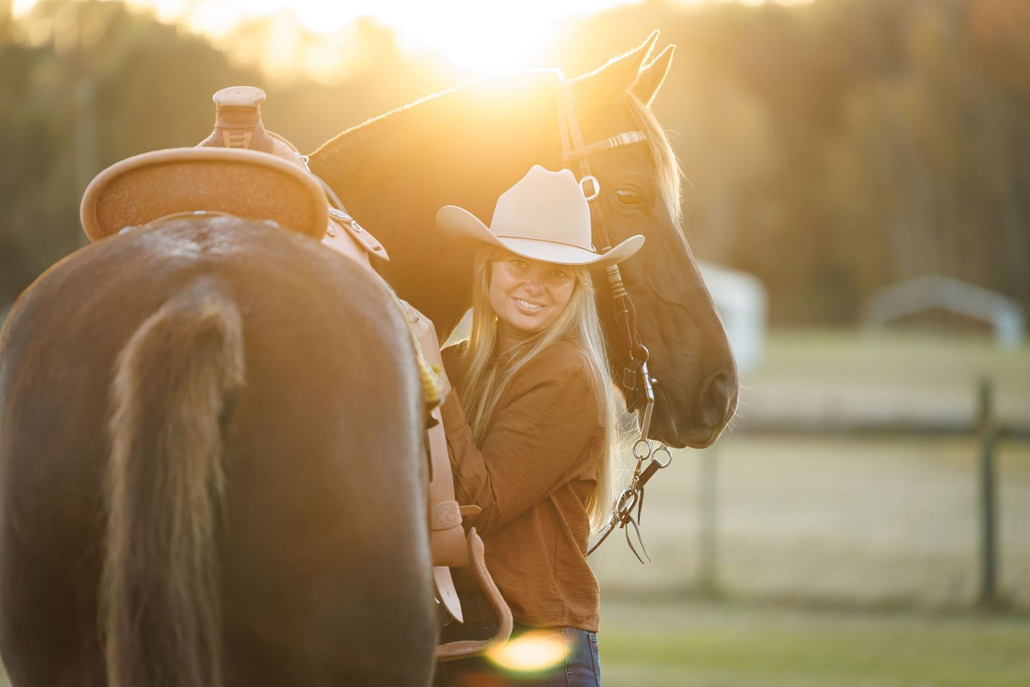 Smiling cowgirl in hat adjusting saddle on chestnut horse in golden sunset light.
