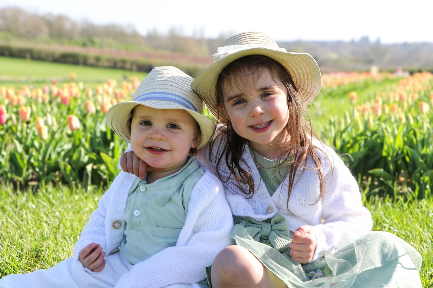 Children sit together in a grassy field wearing sun hats and light colored clothing on a bright spring day.