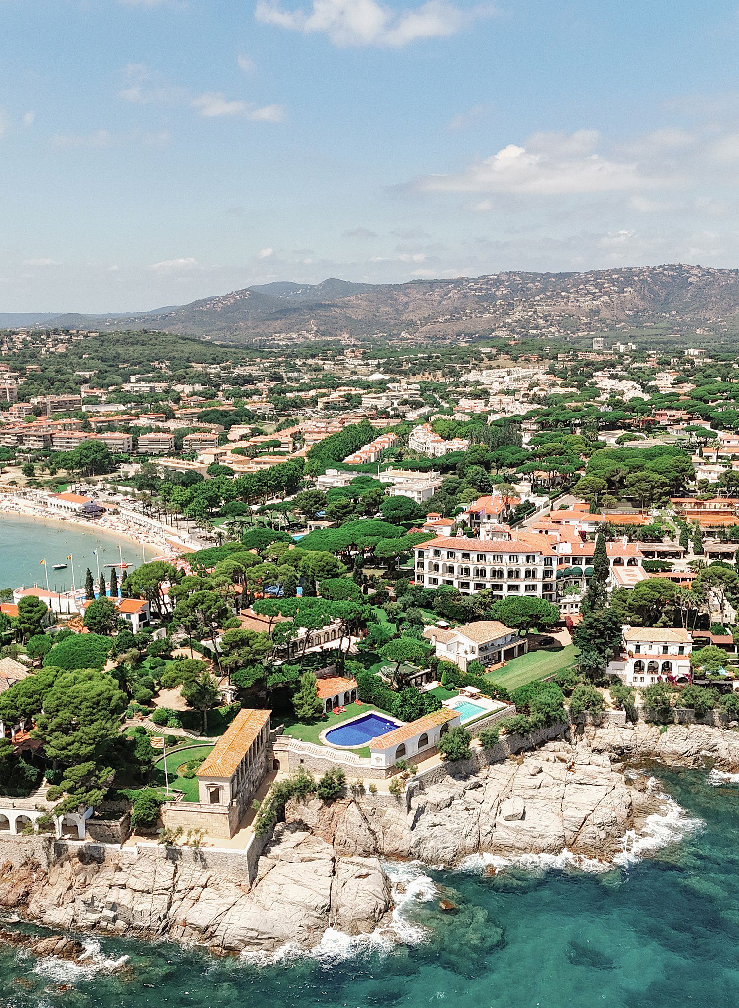 Aerial view of a Mediterranean coastal town with luxury villas, swimming pools and rocky cliffs along the turquoise sea.