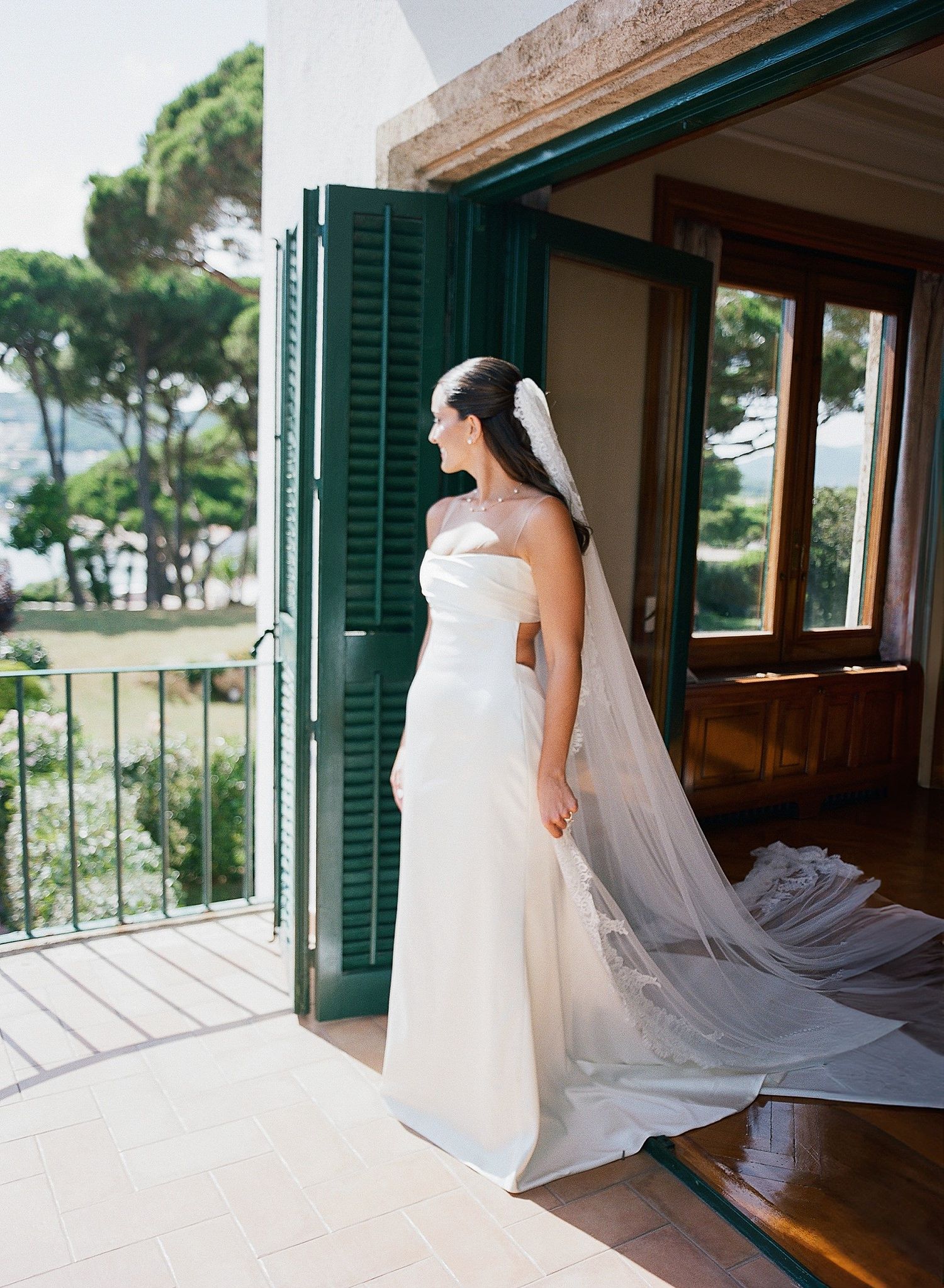 A bride in a flowing white gown with a long veil stands in a rustic room with green shuttered windows and natural light.