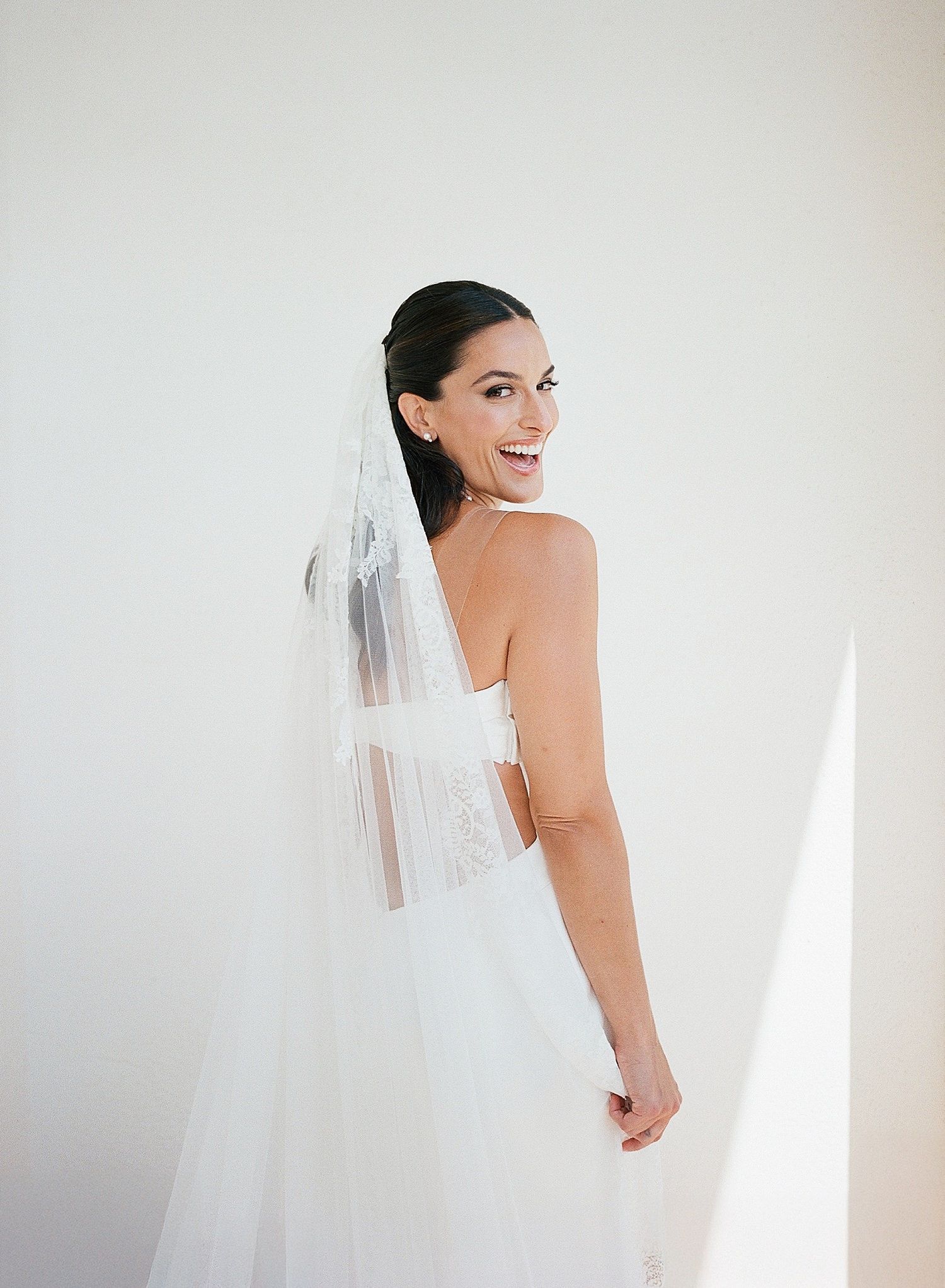 A joyful bride wearing a white wedding gown and veil poses against a light background.