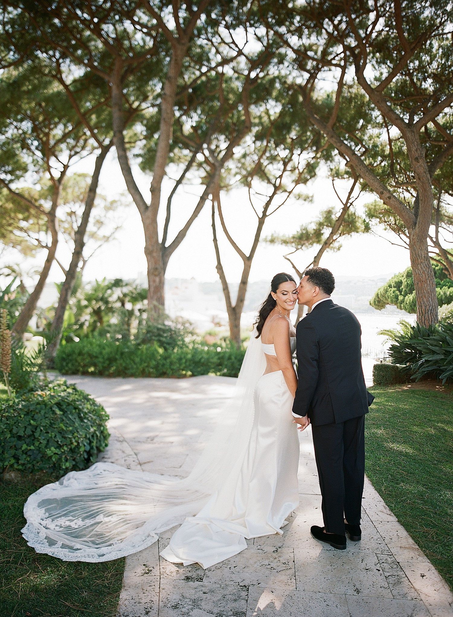 A couple shares an intimate moment on a garden path surrounded by trees, with the bride's long lace veil trailing behind.