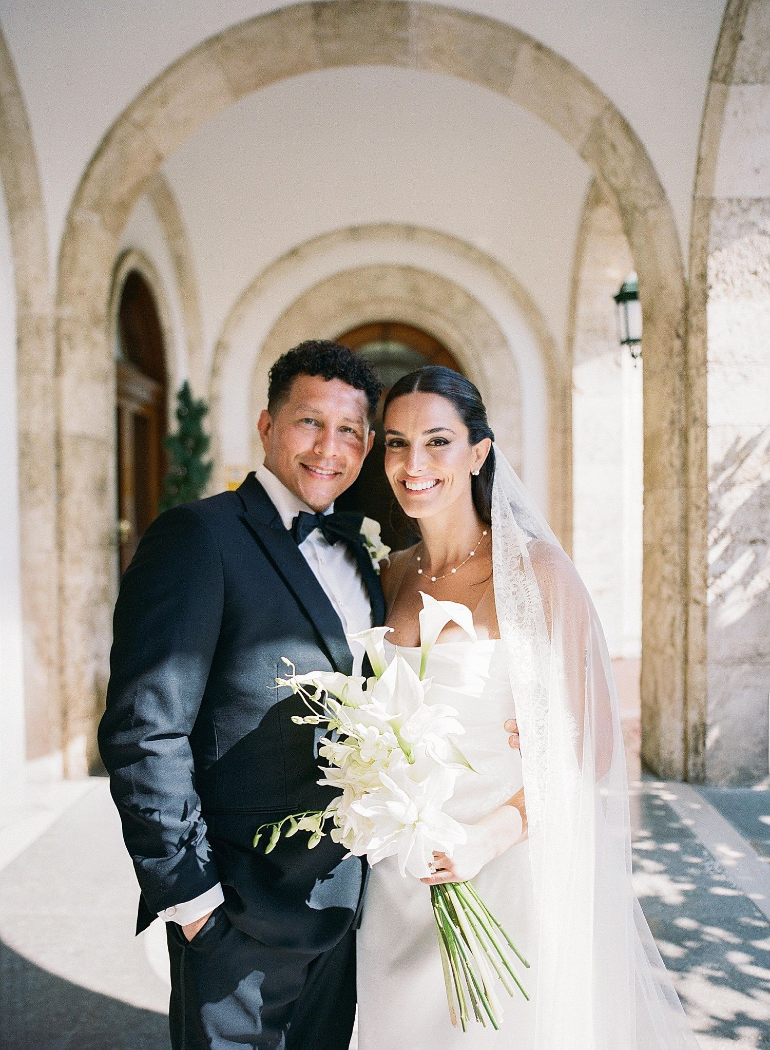A wedding couple shares a joyful moment under elegant stone arches, with the bride holding a white floral bouquet.