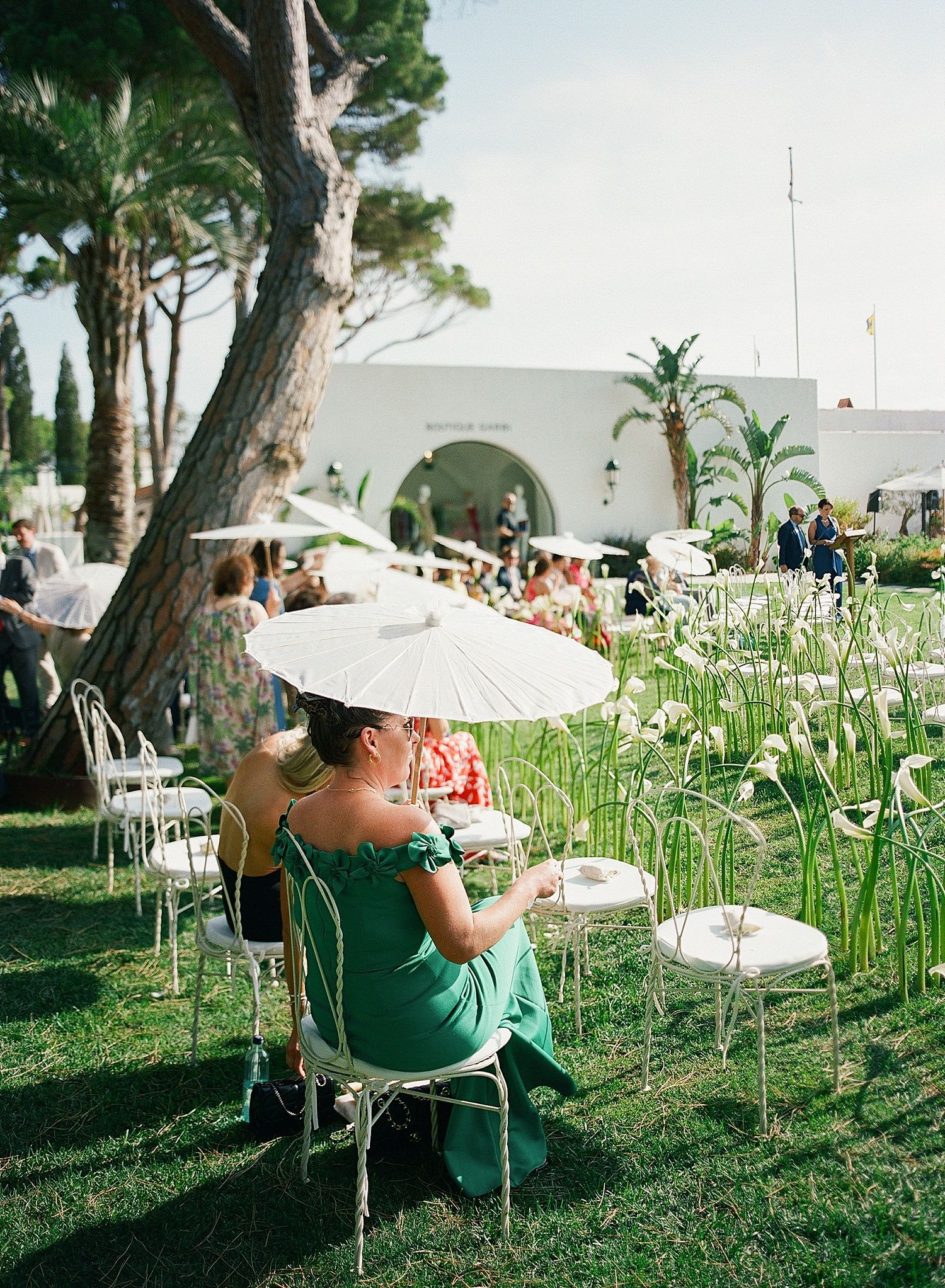 Guests sit in white chairs on a grassy lawn with tall flowers during an outdoor event.