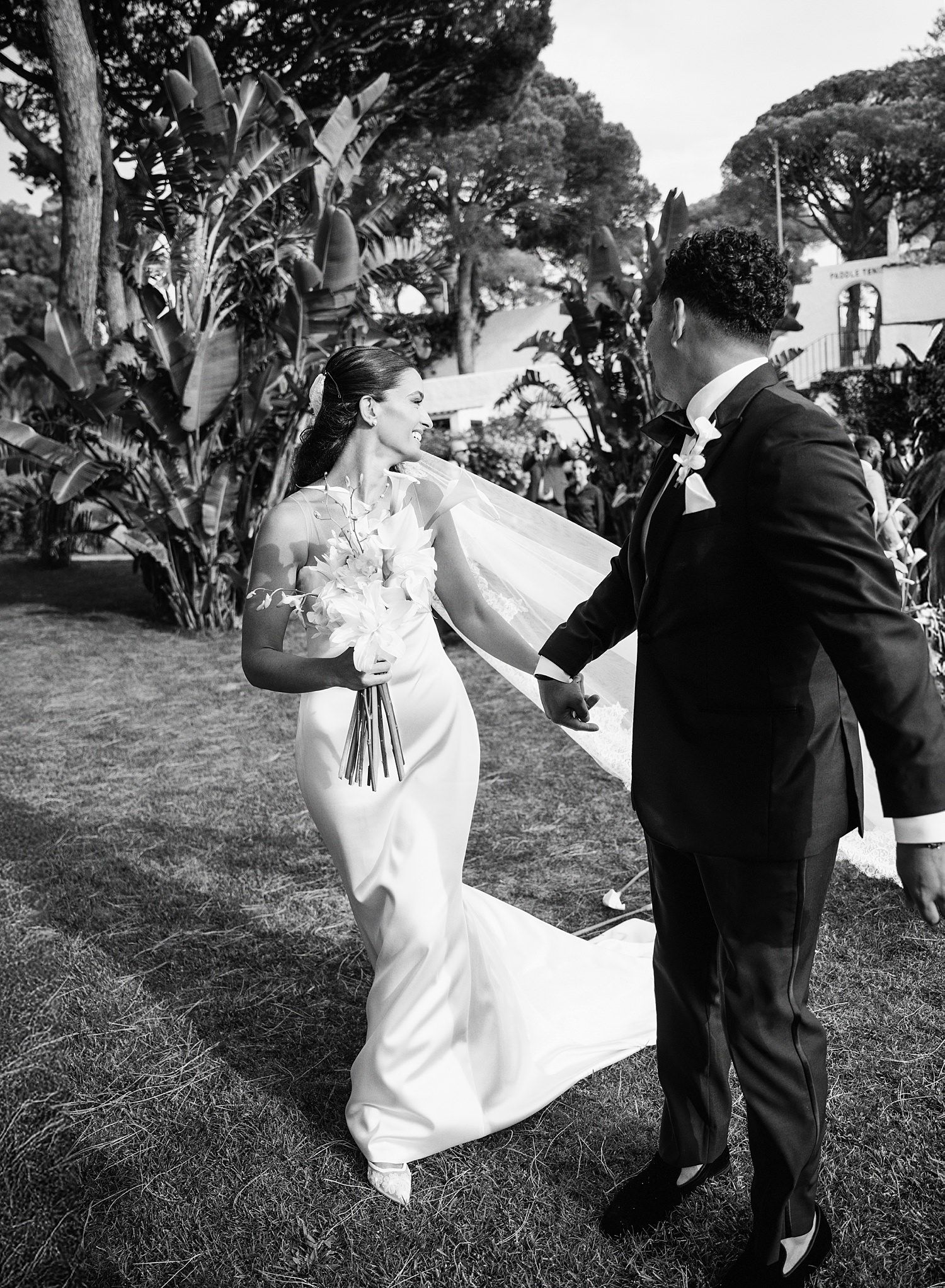 A couple shares a joyful moment during their outdoor wedding celebration, with tropical plants in the background.
