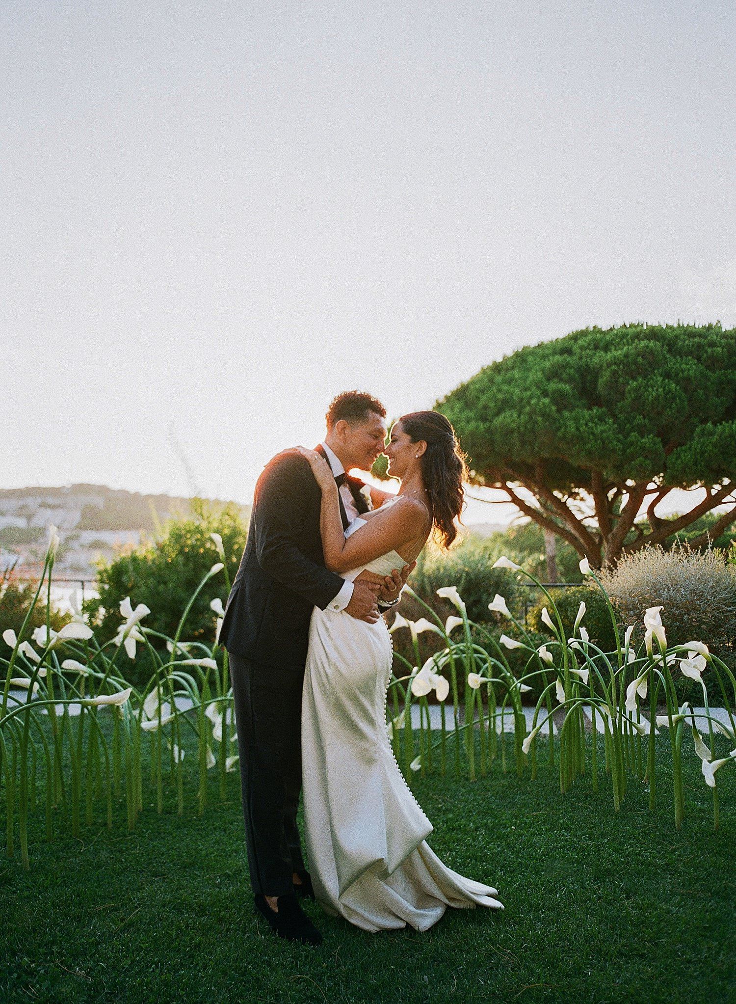 A couple shares a romantic kiss in a garden filled with white calla lilies at sunset by the ocean.