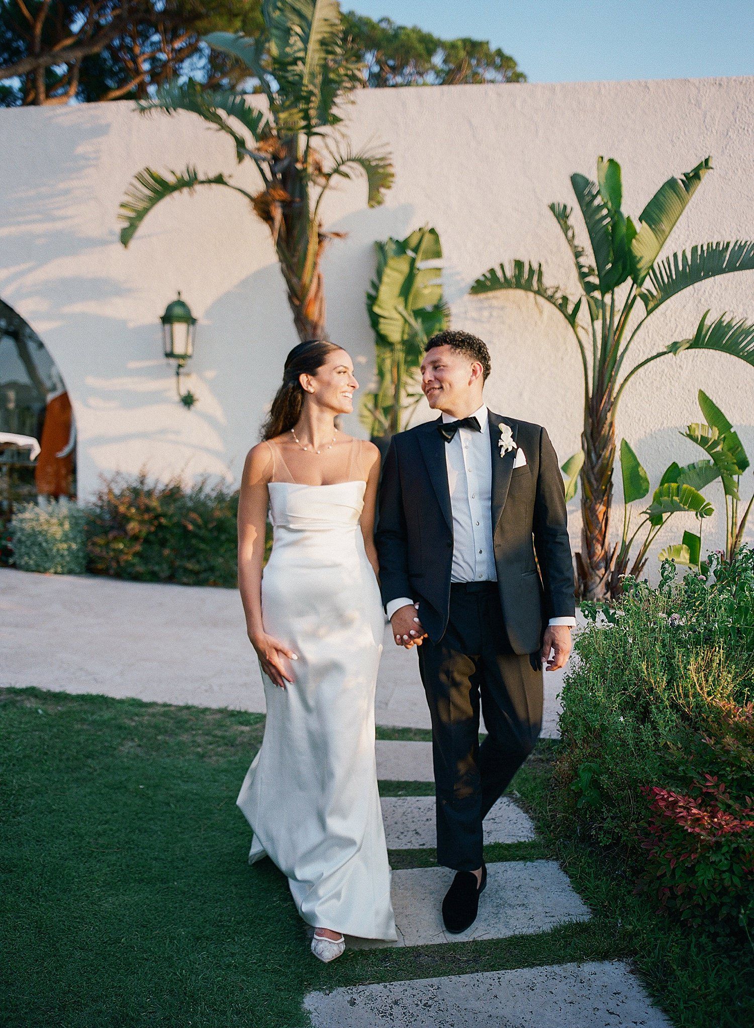 A couple walks together along a garden path with tropical palm trees and flowering plants in an outdoor setting.