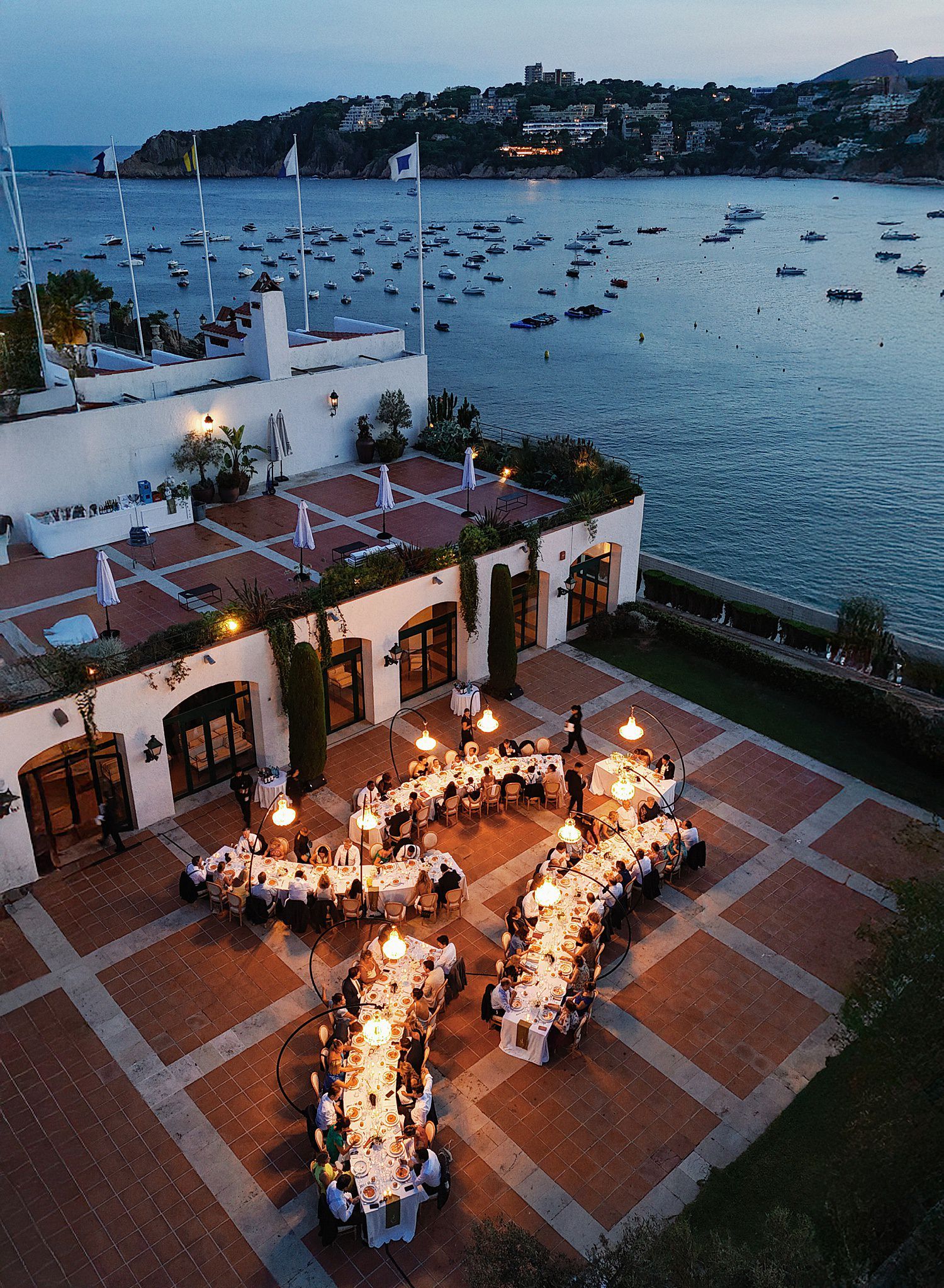 Aerial view of a seaside wedding reception with long tables and string lights on a terrace overlooking a harbor at dusk.