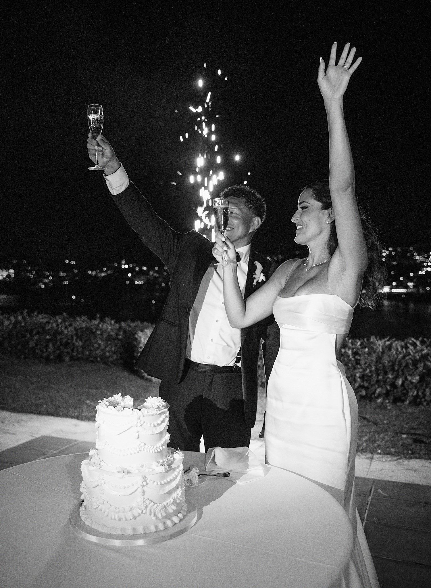 A couple celebrates at their wedding reception with raised arms next to a white tiered cake, captured in black and white.