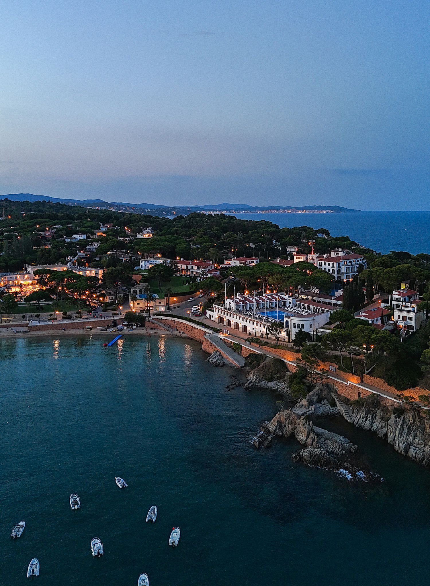 Aerial panoramic view of a Mediterranean coastal town with white buildings and boats anchored in the blue bay at dusk.