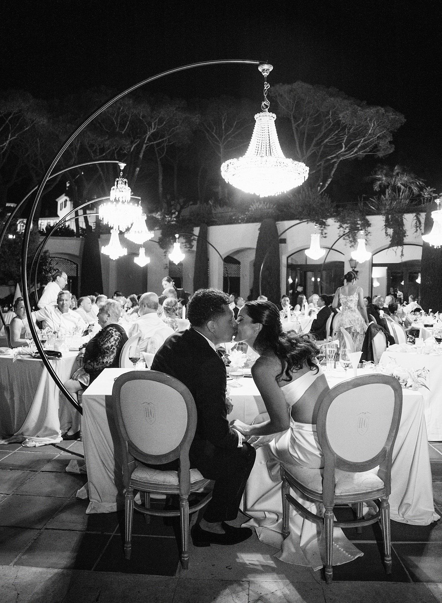Wedding reception guests seated at elegant tables under crystal chandeliers in a dimly lit outdoor venue at night.