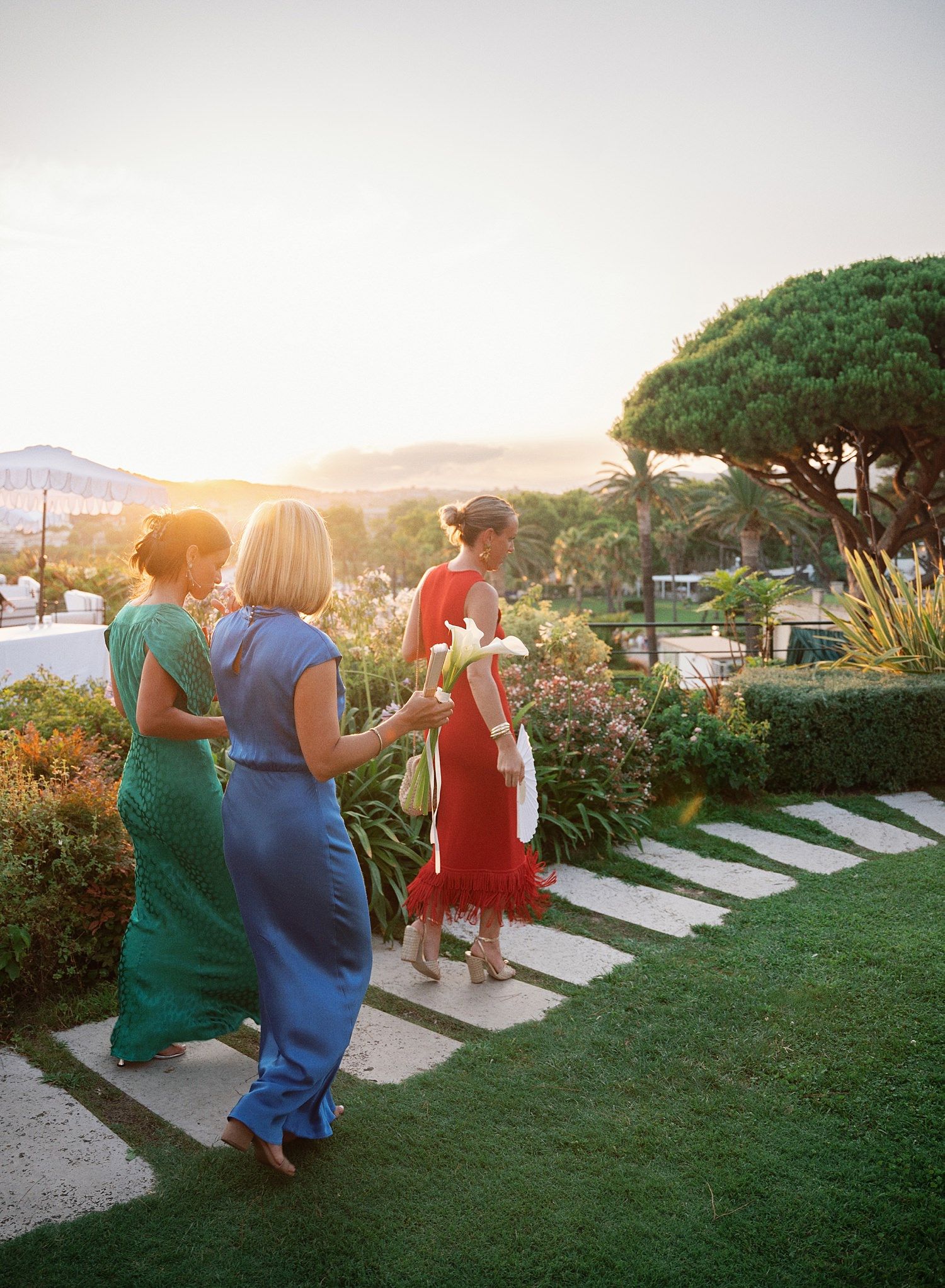 People walking on stone path through garden at sunset with warm golden light filtering through trees and landscaping.