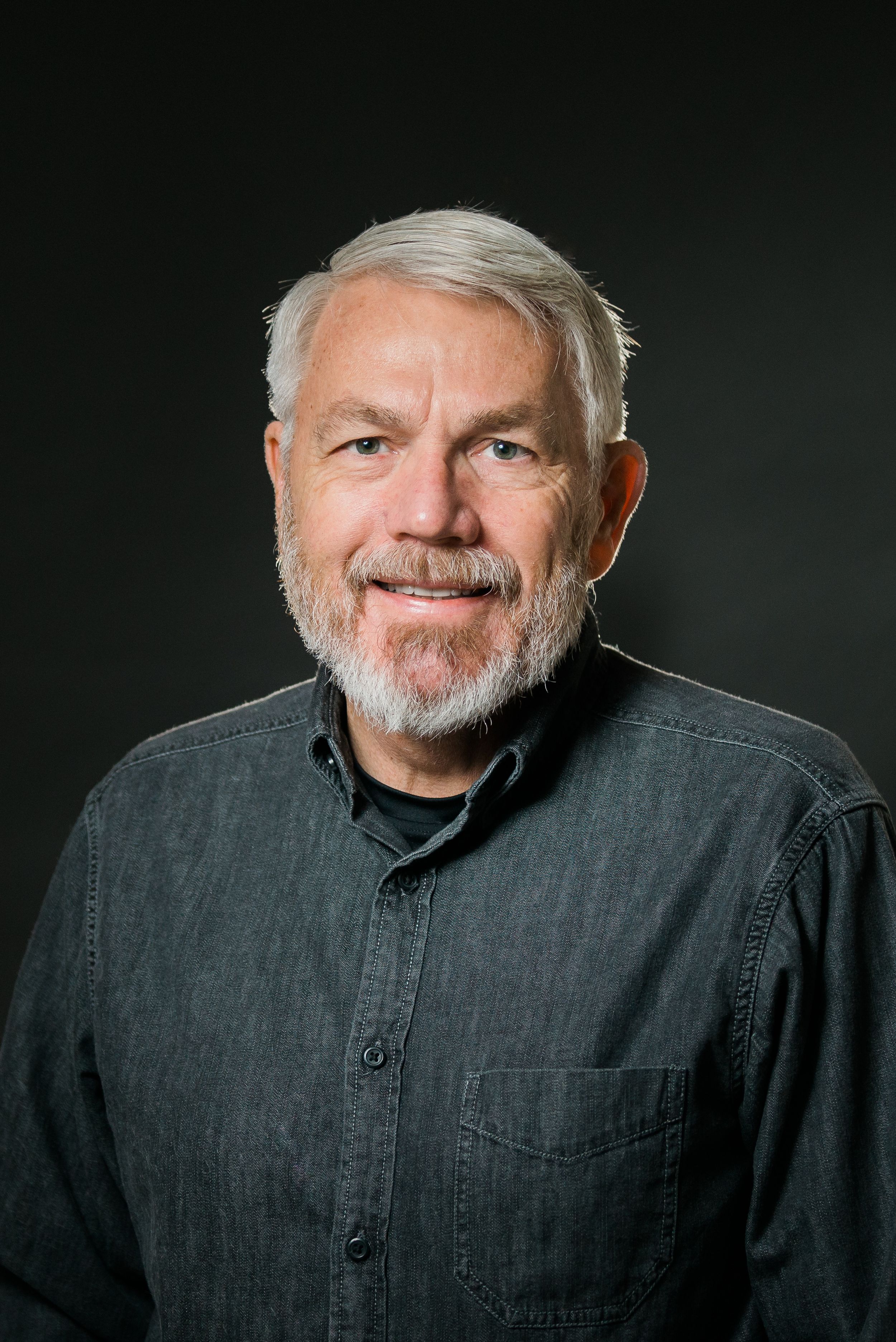 Studio portraits of person with grey beard wearing dark button-up shirt against black background.